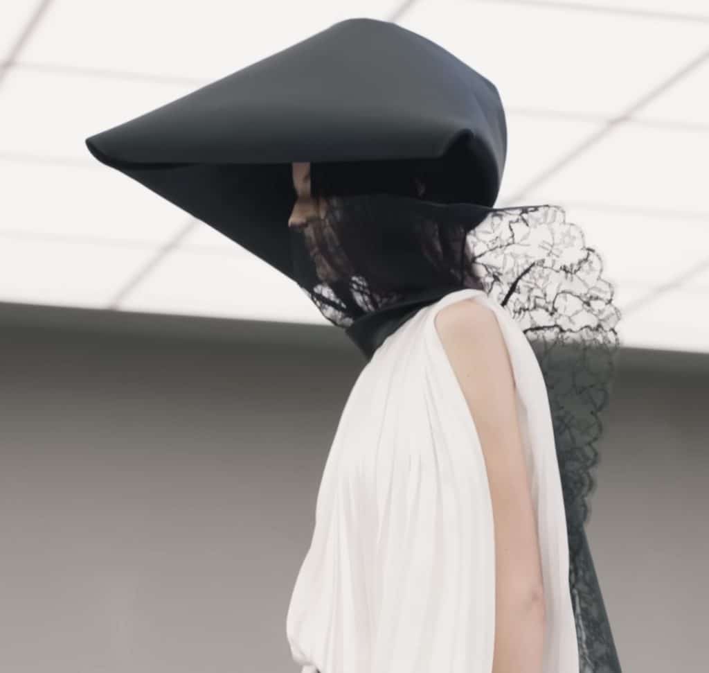 A model with a pointed hat walks down the runway, with the side view of the lace scarf showing.