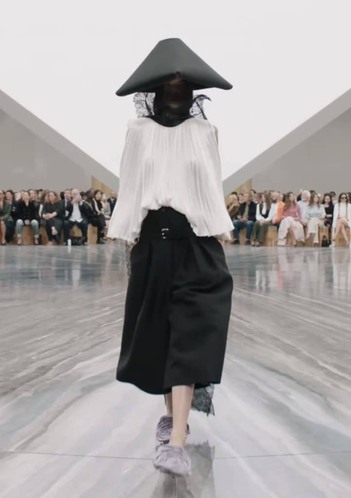Image of model walking the runway in a pleated, flowing silk blouse with baggy black pants. The model is also wearing a pointed black hat, with a piece of black lace that wraps around her neck and flows to the back of the garment.