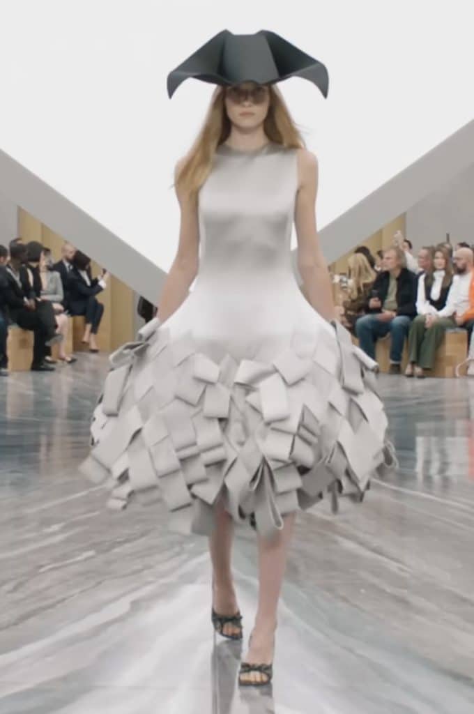 A model in a grey dress walks the runway in a pointed black hat. The dress is fitted on the bust and waist area, then becomes wide from the waist down. The bottom half of the dress has layers of fabric woven together to create a geometric pattern.