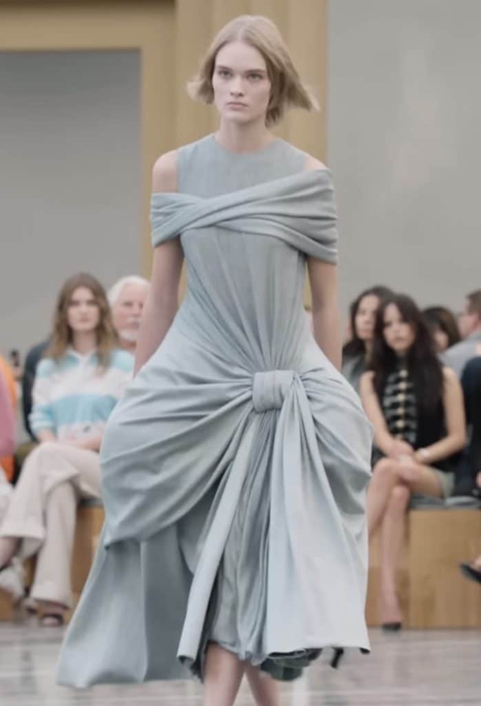The model walks down the runway in a grey dress which is made abstractly. The bust and the waist are slimmed, and then the hips become wider and protrude from the rest of the body creating an interesting shape.