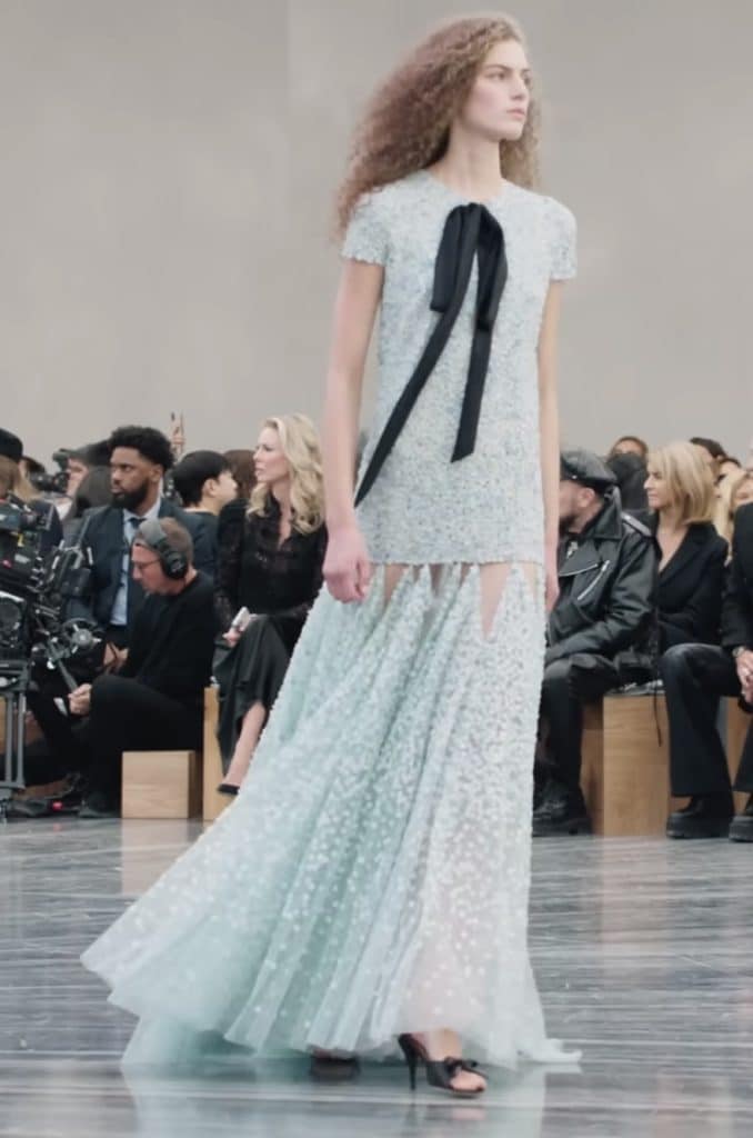 The model walks down the runway in a light blue dress made of mesh, which is slim fitted from the bust to the hips, and then starts to flow from the hips down to the ankles.