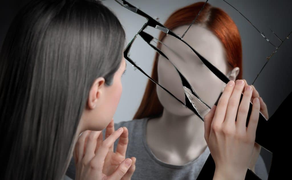 Image of a woman staring at a reflection of herself in a shattered mirror, except it is distorted, deathly pale, missing a face, and has a different hair colour. Meant to convey distorted self-perception.