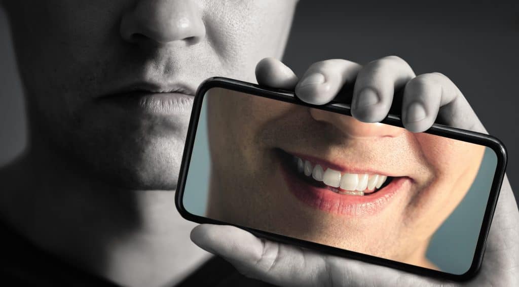 Image of a man frowning in black and white, as he holds up a coloured image on his phone of his lower face, where he is smiling.