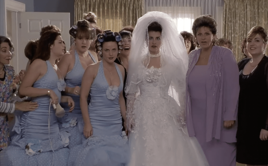 A woman in a wedding dress stares into the camera surrounded by her bridesmaids