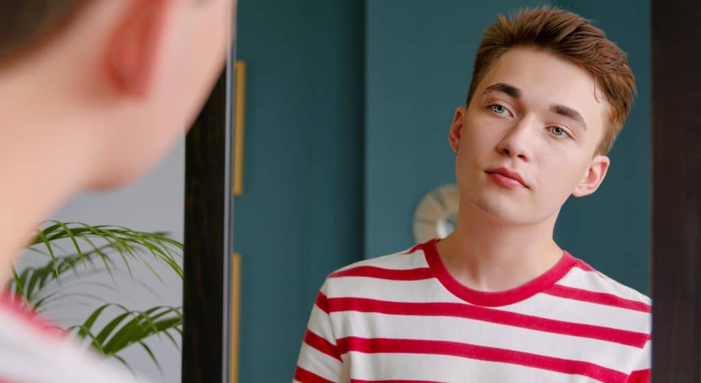 A young teenage guy looks in the mirror at his reflection. Pure beautiful skin and natural beauty of a young man. Selective focus.