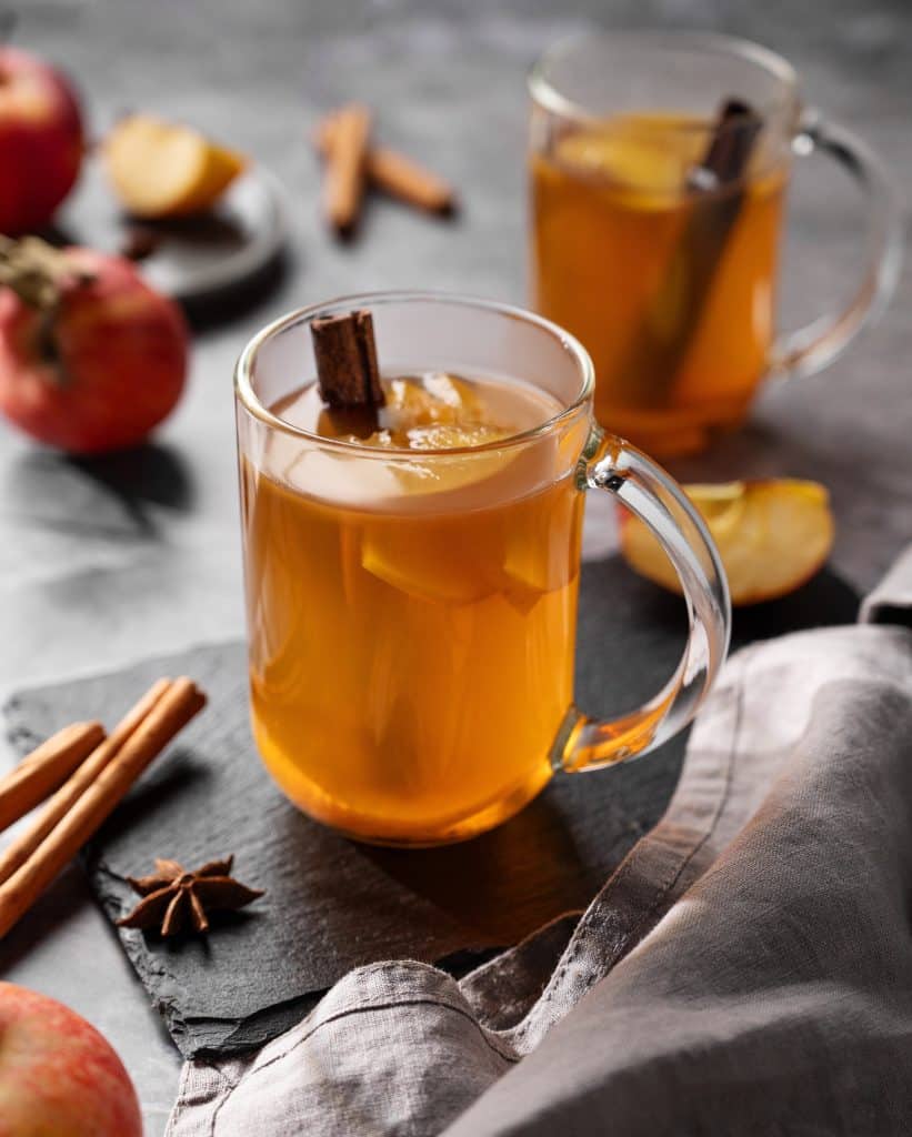 A clear glass of apple cider with a cinnamon stick sits on a table.