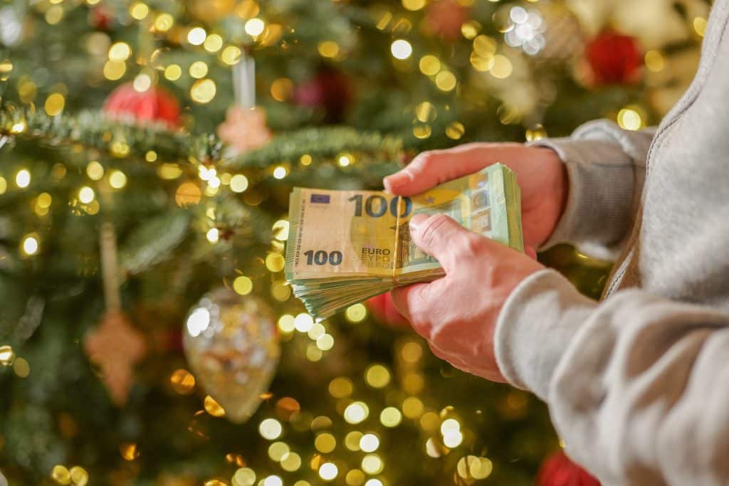A person holds a stack of Euros in front of a Christmas tree.