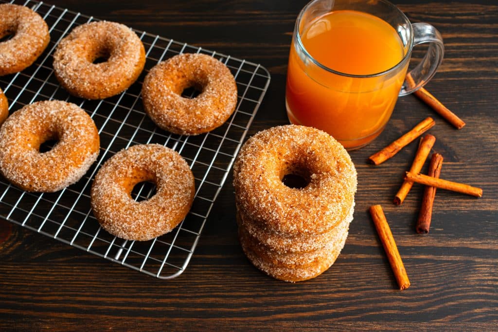 A tray of apple cider donuts sits next to a glass of apple cider and cinnamon sticks.