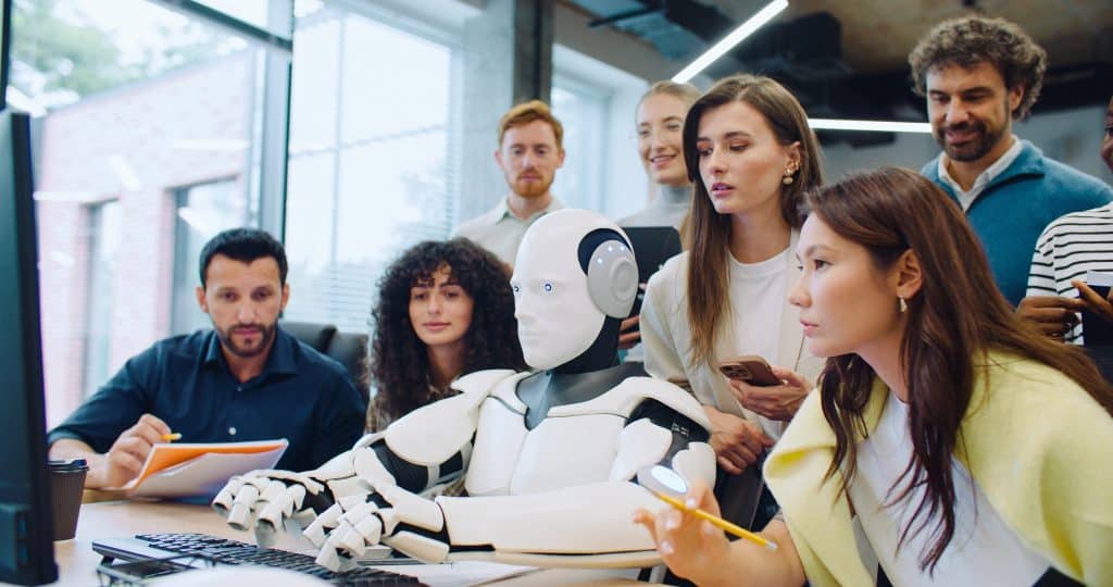 A group of office workers gathered around a robot typing on a computer.