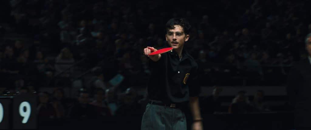 Timotheé Chalamet holding a paddle while pointing at something off screen in 'Marty Supreme.'