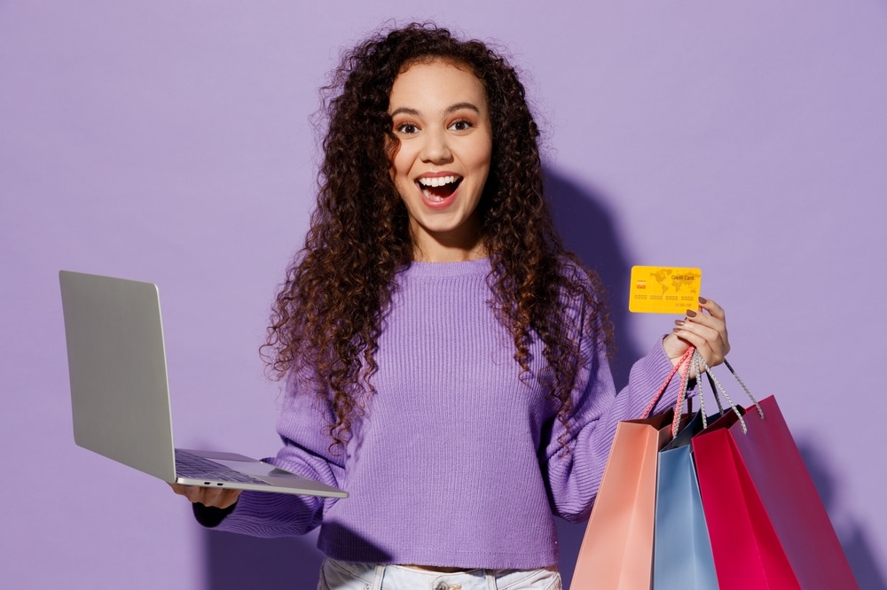 Young fun IT woman wear pullover hold paper package bag after shopping use work on laptop pc computer use credit card isolated on plain pastel light purple background Black Friday sale buy day concept.