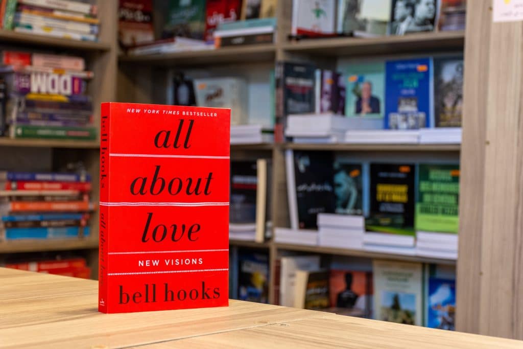 A red copy of the book All About Love by bell hooks stands on a table in front of a book case.