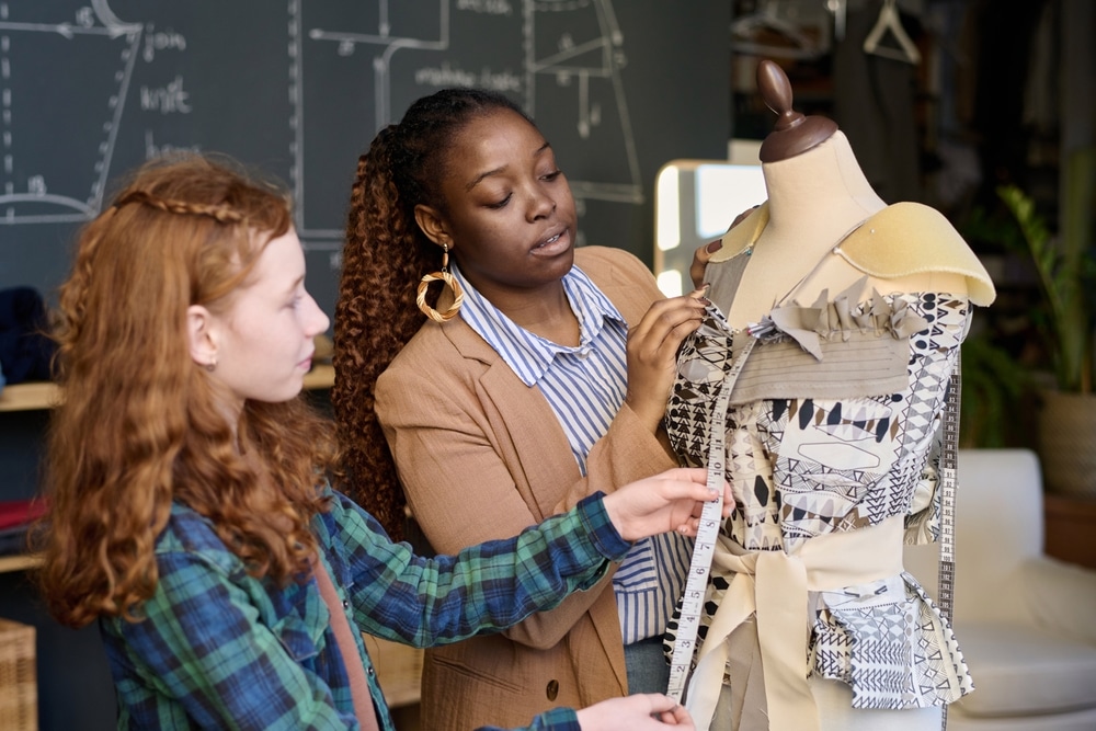 Waist up portrait of teenage girl with female teacher fitting designer clothes to mannequin in atelier copy space.