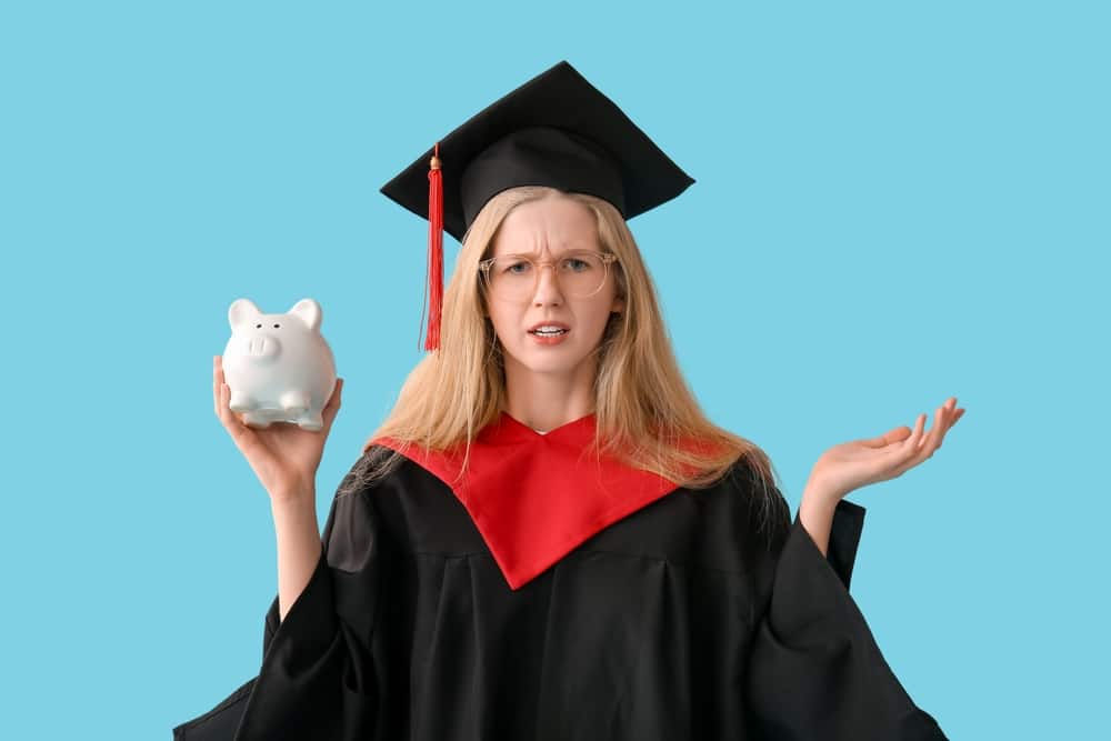 Female student in graduation cap and gown holding a piggy bank while shrugging.