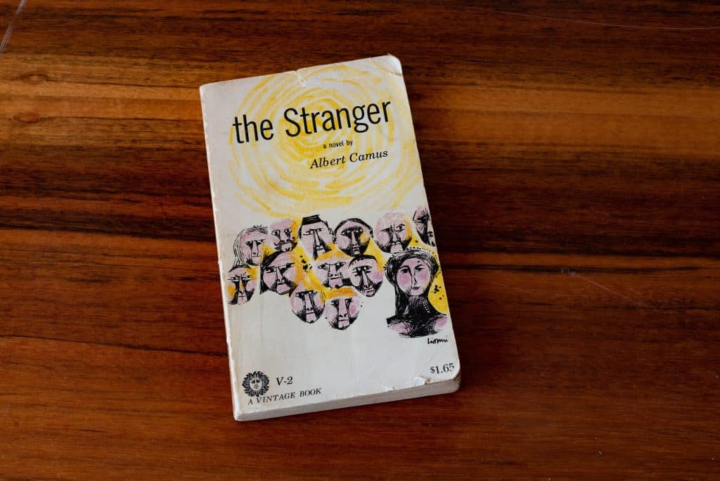 A copy of The Stranger by Albert Camus lies on top of a table, slightly worn. The cover is white and yellow with distorted faces on it.