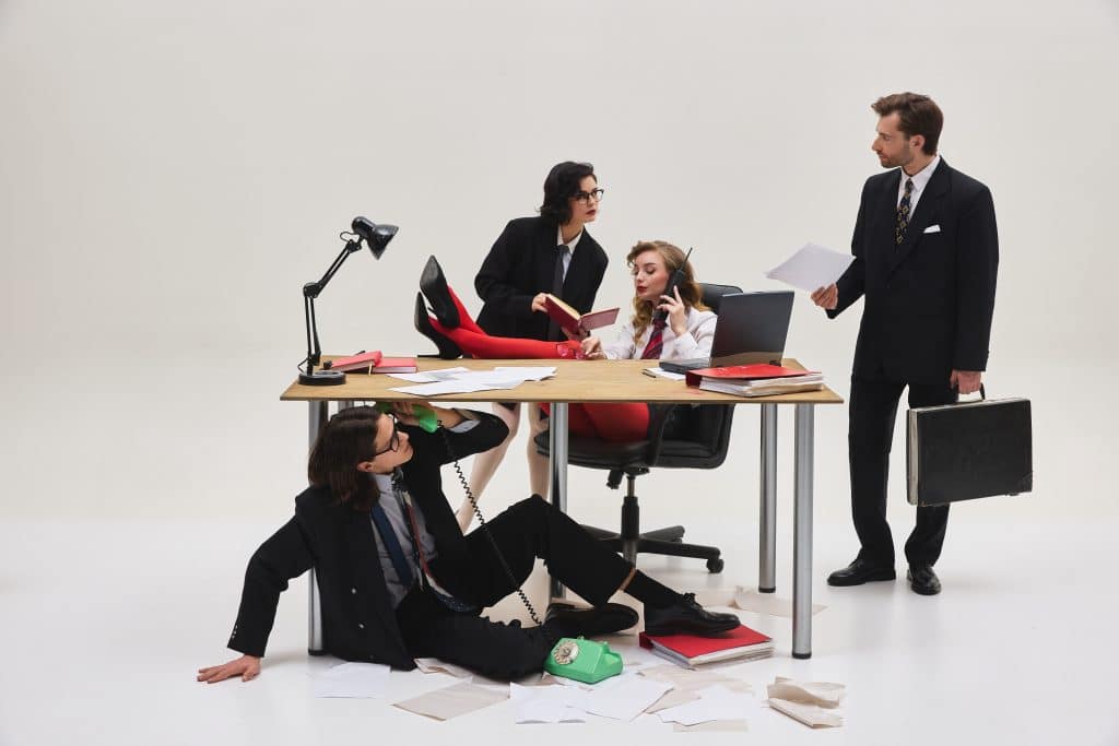 Four office workers in business attire crowd around a cluttered desk. One woman sits at the desk on the phone with papers and folders scattered around, while another leans over her holding a notebook. A third person sits on the floor using a rotary phone, and a man in a suit stands nearby holding documents and a briefcase. The scene appears chaotic and humorous, suggesting a hectic workplace.