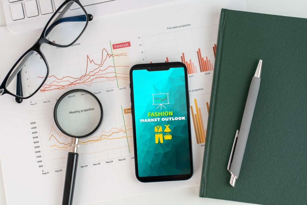 A smartphone displaying Fashion Market Outlook report is placed atop a stack of papers with graphs and charts, accompanied by yellow notebook, keyboard, glasses and magnifying glass.