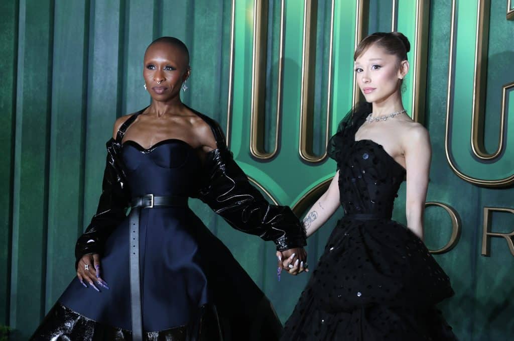 Cynthia Erivo and Ariana Grande stand in black dresses against a green background for promotion of Wicked: For Good.