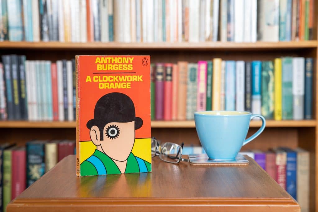 A copy of Anthony Burgess's A Clockwork Orange is propped up in front of a bookcase. A blue mug is to the right. The cover of the book depicts a cartoon of a man with one swirly eye wearing a bowler hat.