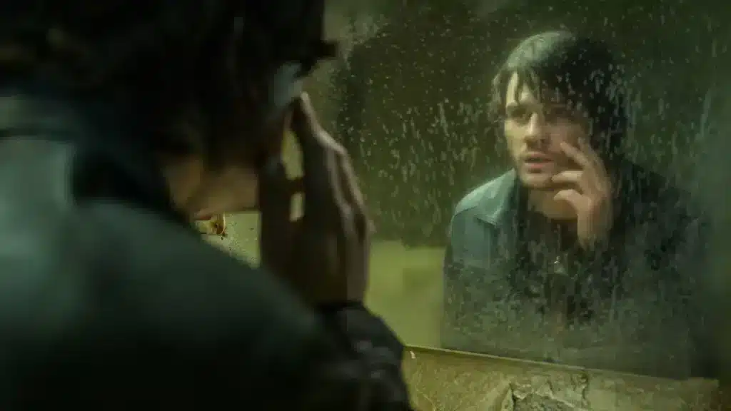 A man stares at himself in a dirty mirror, touching his cheek.