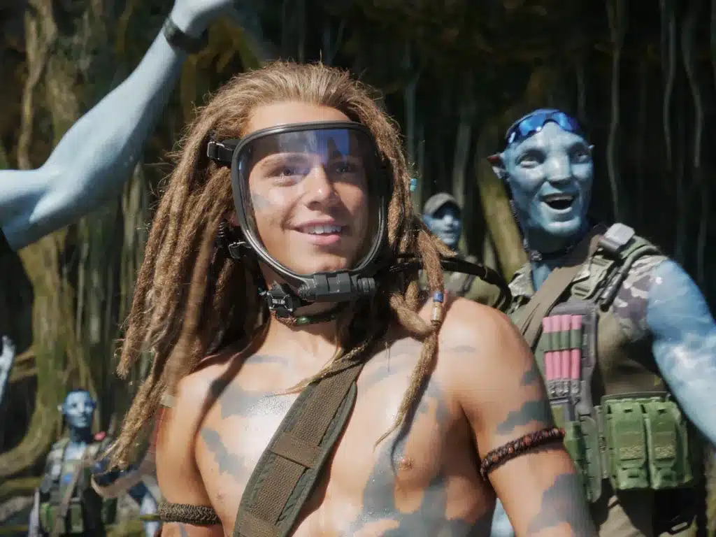 The image shows a teenage boy with a sci-fi mask over her face. Behind him stands a blue Na'vi Avatar.