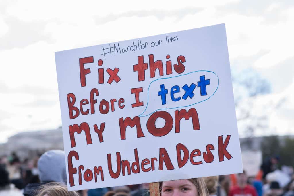 Image shows protestors at a March For Our Lives rally holding up a sign that says '#Marchforourlives - Fix this Before I text my mom from Under A Desk.'