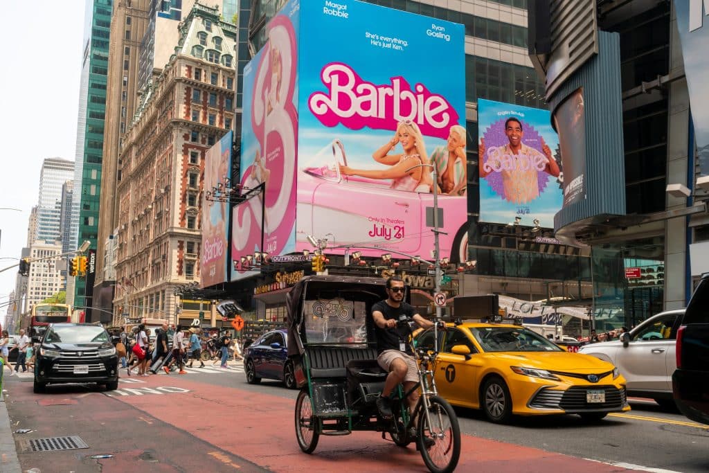A shot a busy city street filled with cars and people. Looming over them is a massive Barbie advertisement featuring Barbie in her car with Ken in the backseat.