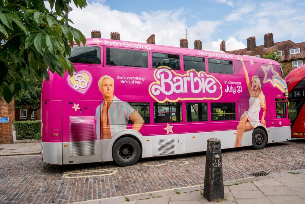 A bus covered in Barbie advertising is parked on a brick road.