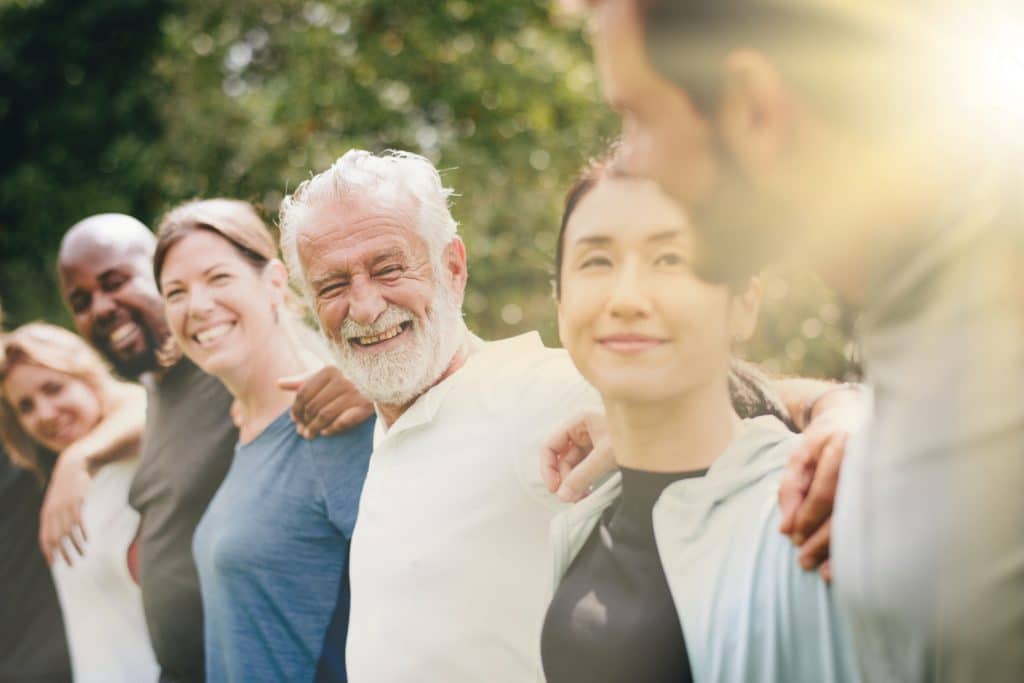 Diverse group of people standing together outdoors, smiling and embracing. Sunlight filters through trees, creating a warm, friendly atmosphere of unity and happiness. Friendship and unity image.