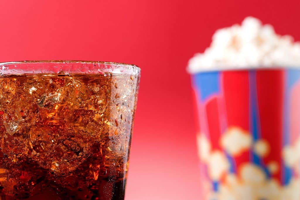 A close up shot of a glass of brown soda and a blurry popcorn bucket in the background.