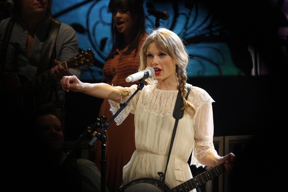 Taylor Swift points to the crowd with her right hand and sings into a microphone, holding her instrument