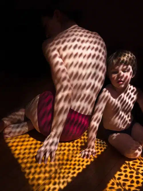 A full color photograph of two young boys, half naked, sitting in front of a shaded window, half in shadows.