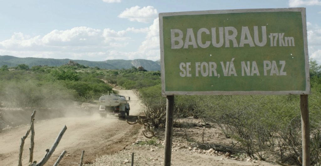 Truck traveling a dirt road with a sign in the foreground saying "Bacurau – 17 km – If you're going, go in peace" (translated).