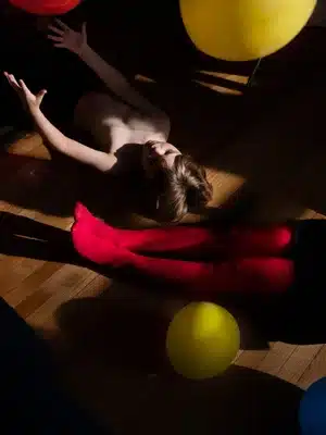 A full color photo of a shirtless boy laying on the ground, arms outstretched. Next to the boy are a pair of legs laying diagonally on the ground.