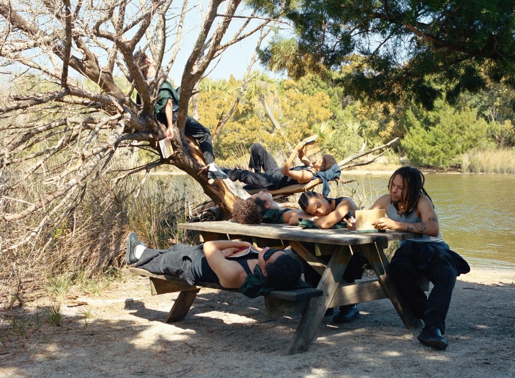 A full color photograph of males sitting on a tree, sitting at a picnic table by a body of water.