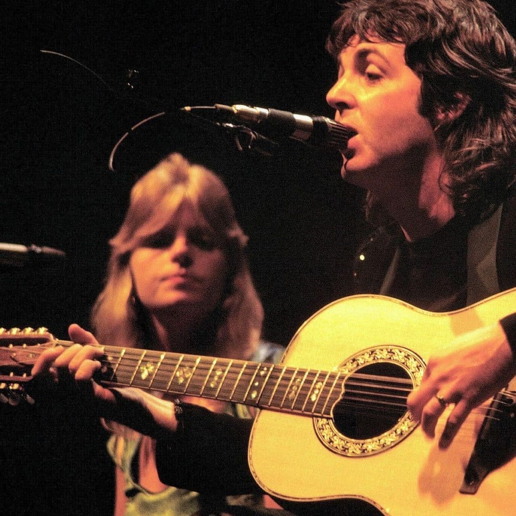 Linda (left) and Paul (right) sing on stage. Paul is playing acoustic guitar and singing into the microphone; Linda is looking at him.