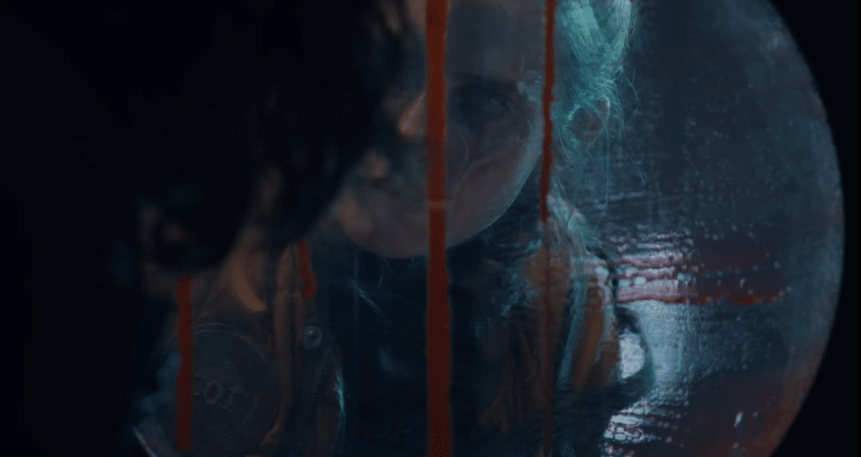 Image pf Ava looking in through the submarine's window, which has blood slowly running down the outside of it. The left side of her face and her eye are visibly scared. It makes for a conventionally unsettling picture.