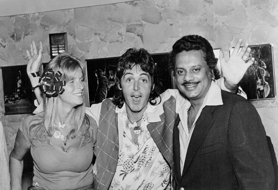Linda (left), Paul (center), and Bhaskar Menon (right) stand facing the camera, Paul embracing them both; Linda looking to the right.