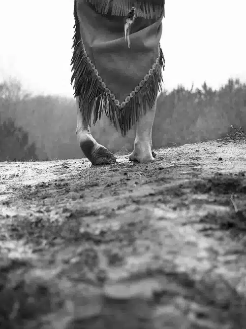 Black and white photograph of a person from the waist down wearing Native clothing walking away from the camera, feet cracked and dirty.