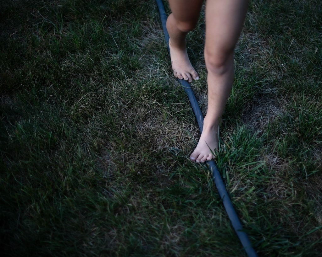 A full color photograph of a pair bare feet and legs in the grass, walking on a garden hose.