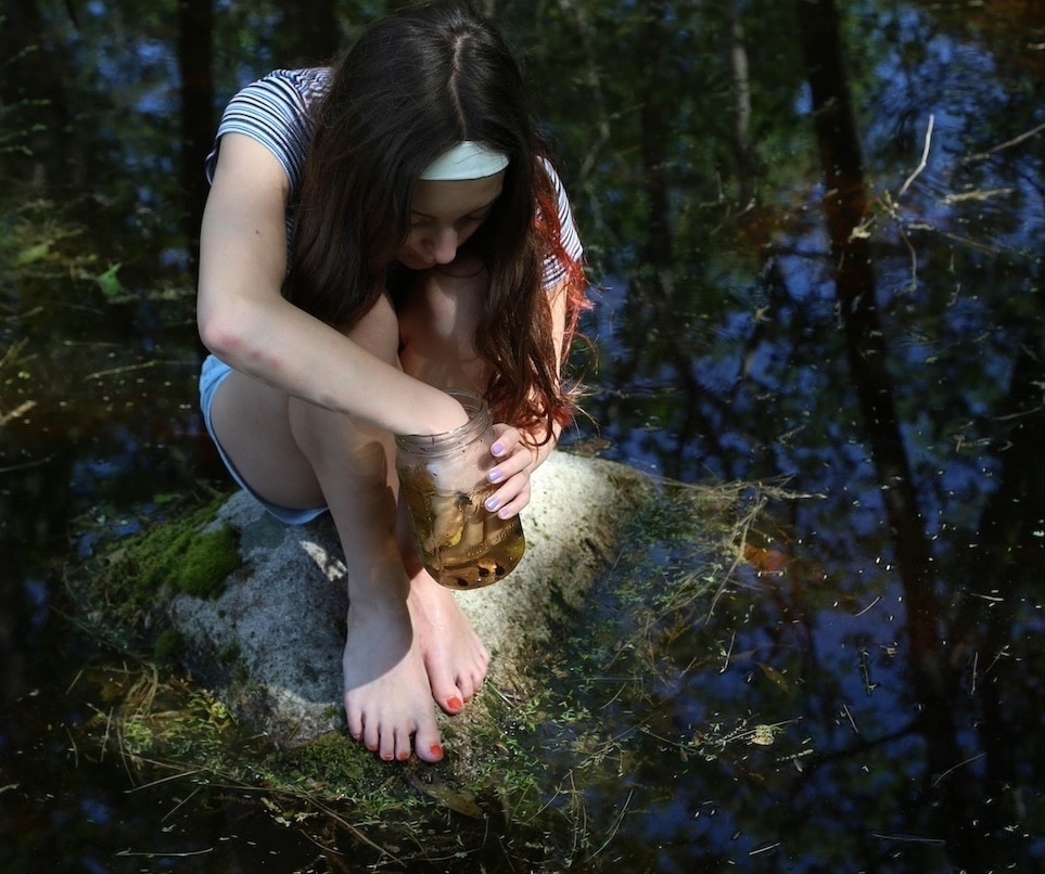 A photograph of a young girl crouching in the woods, surrounded by flowers and trees, with her hand in a glowing jar.
