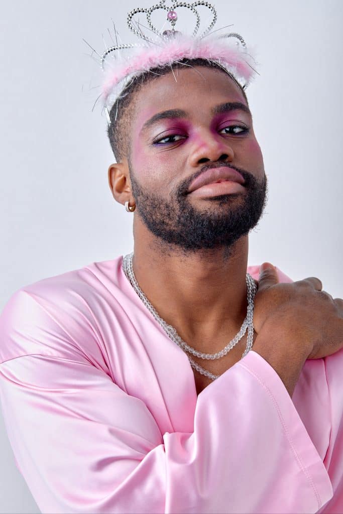 Image of a black masculine looking man with a beard, in a pink shirt with a clear beaded necklace, a lobe piercing and a fluffy pink tiara. He has some very pretty dark pink eye shadow on, which fades to a white shimmer at the outer corners of his eyes, which are underlined with a blue toned purple. The man is conventionally attractive.