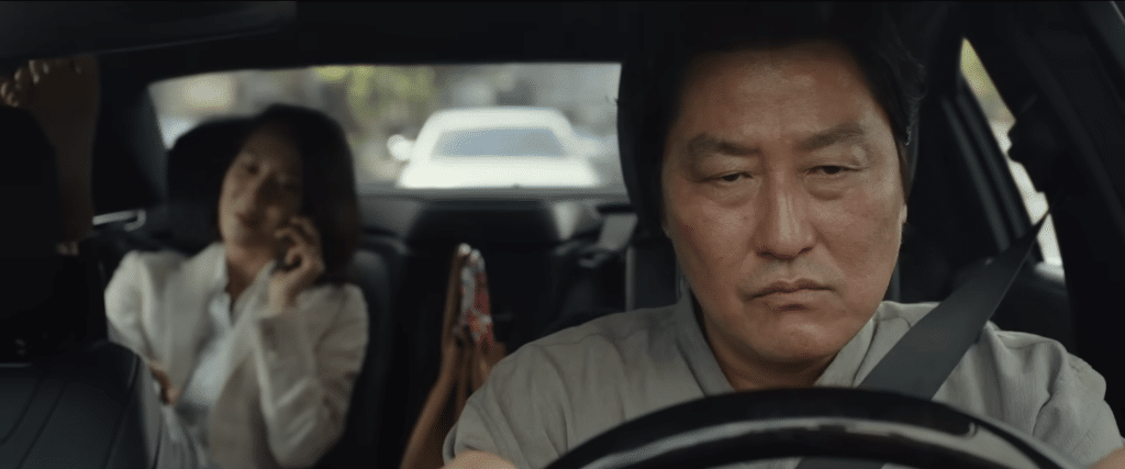 Song Kang-ho as his character Kim Ki-taek, driving with a disgruntled face. Cho Yeo-jeong as her character Yeon-Kyo is sitting in the backseat.