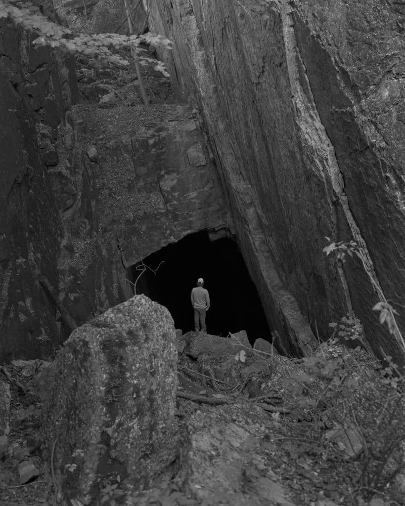 A black and white photograph of the outside of a cave with a man standing in the mouth of it.