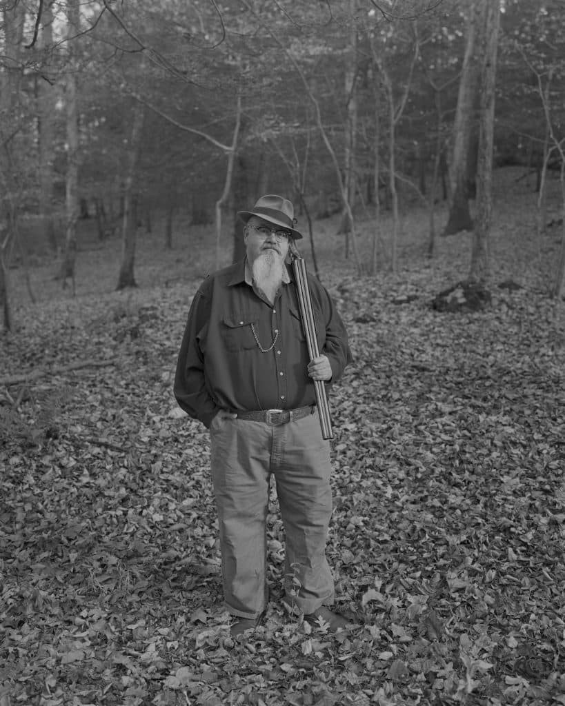A black and white photograph of a man with a long beard standing in the woods with a rifle.