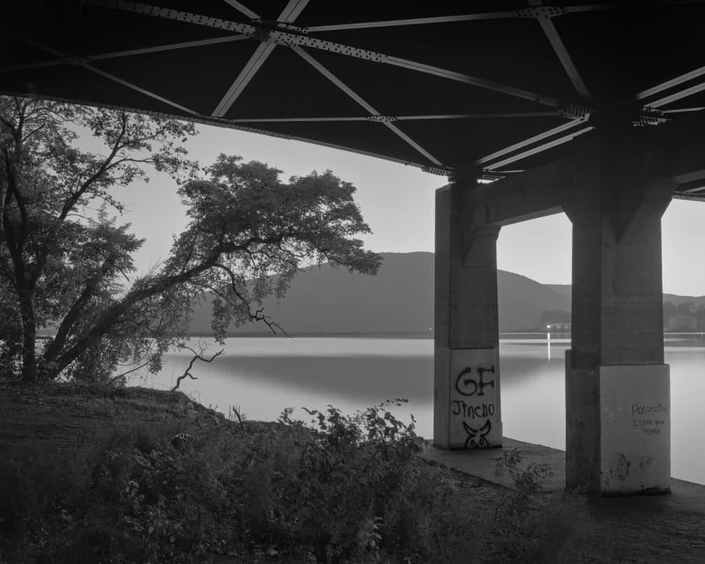 A black and white photograph of the Hudson River under a bridge. Hills can be seen in the background, a tree appears from the side in the midground opposite graffiti on the leg of the bridge, and grass in the foreground.