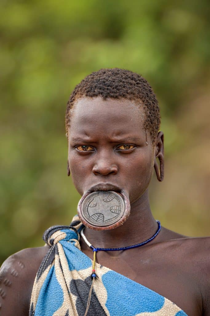 Image of a short-haired black woman with a stretched lower lip and stretched ear lobes staring into the camera. Her lower lip has a clay disc in it. She appears to have some scarification on at least one shoulder.