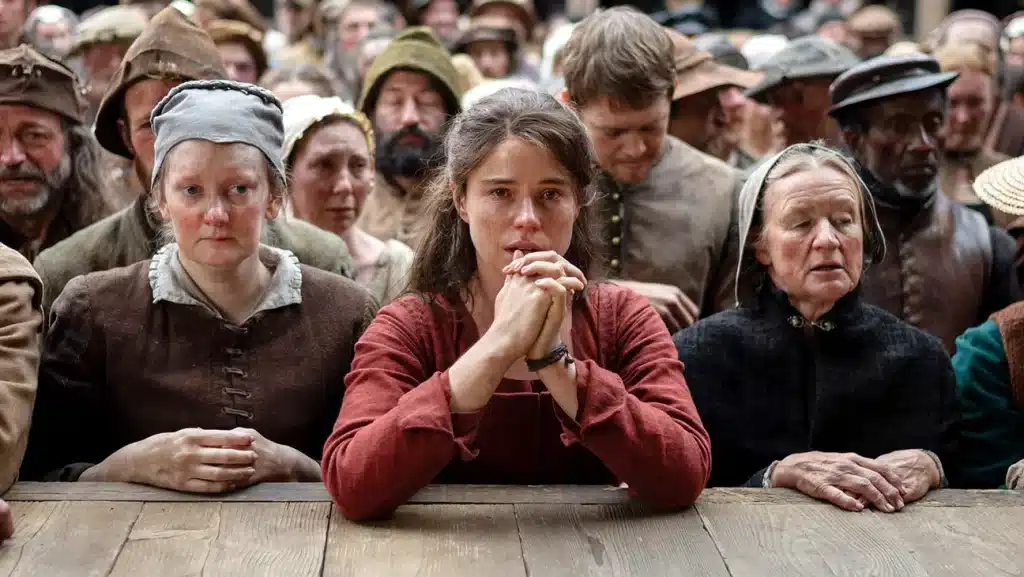 Jessie Buckley praying with hands, staring into the camera. She is looking to the stage.