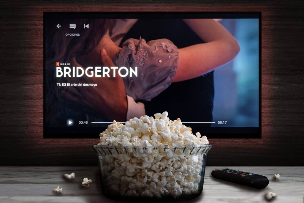 A bowl of popcorn sits on a table next to a remote, in front of a TV screen with Bridgerton playing.
