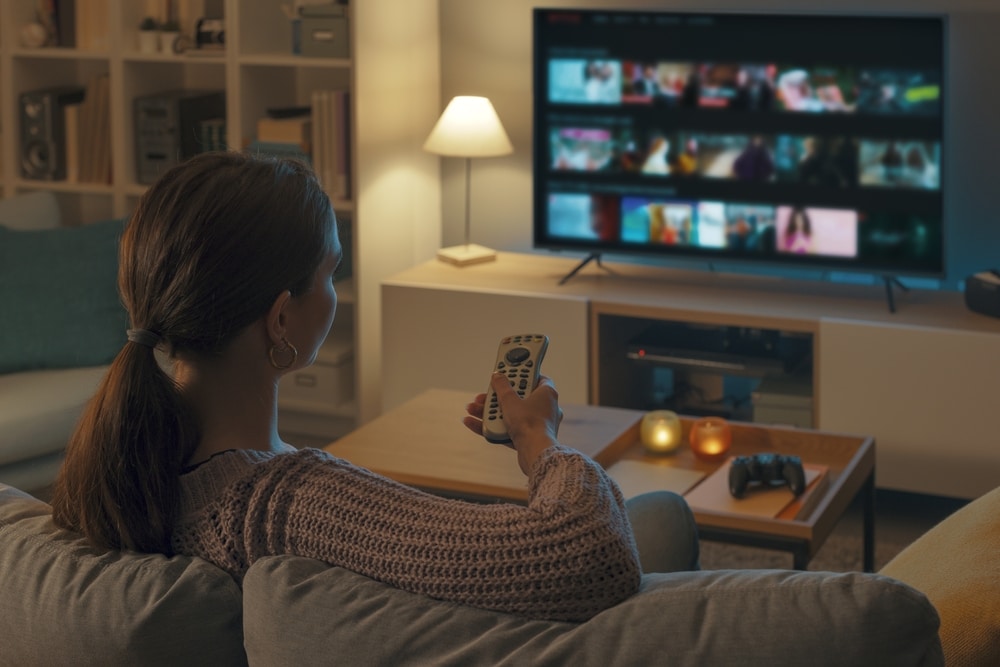Image of woman sitting comfortably on a couch with a remote in her hand pointed towards a TV.